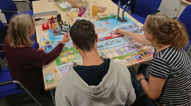 Drei Personen sitzen an einem Tisch und sind in ein Brettspiel vertieft.